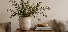 A ceramic vase of leafy branches with a stack of books on a wooden table